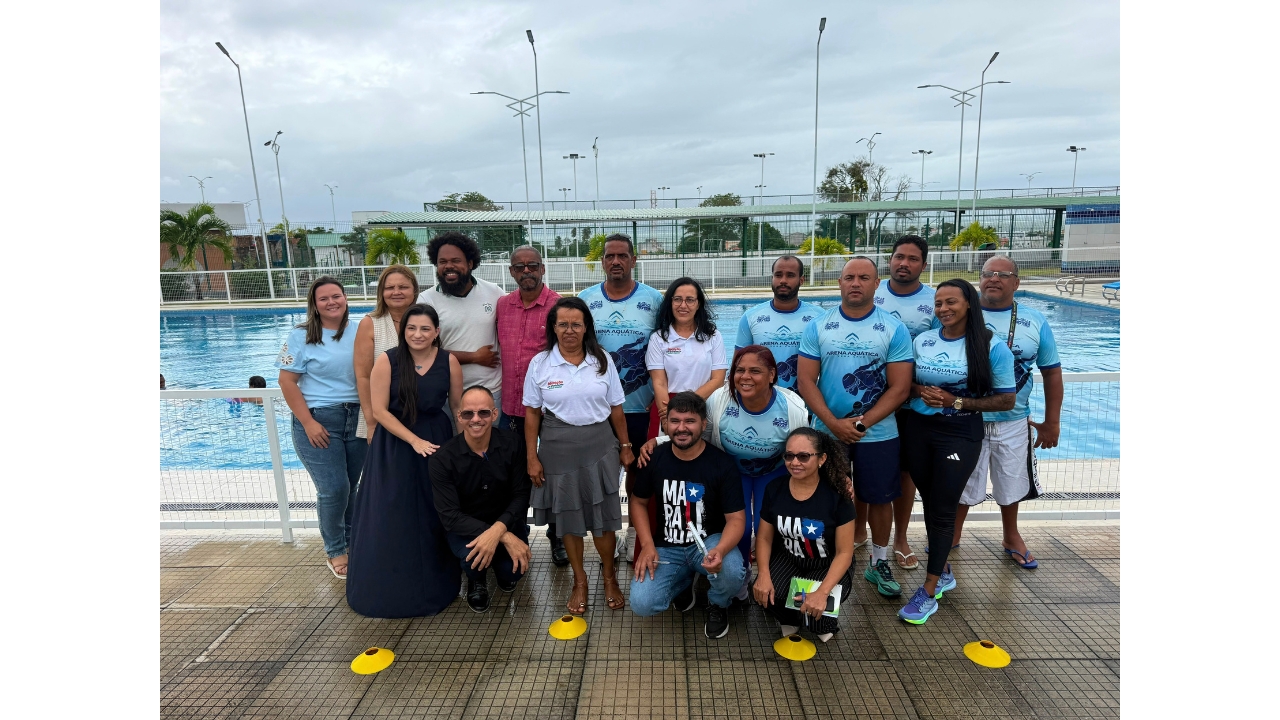 Grupo multirracial de cerca de 17 adultos posa em uma área externa junto a uma piscina. Parte deles usa camisetas azuis com a inscrição “Arena Aquática”, enquanto os demais vestem roupas casuais. Ao redor, é possível observar postes de iluminação, árvores e coqueiros. Atrás do grupo, um portão baixo de ferro branco separa a área da piscina.