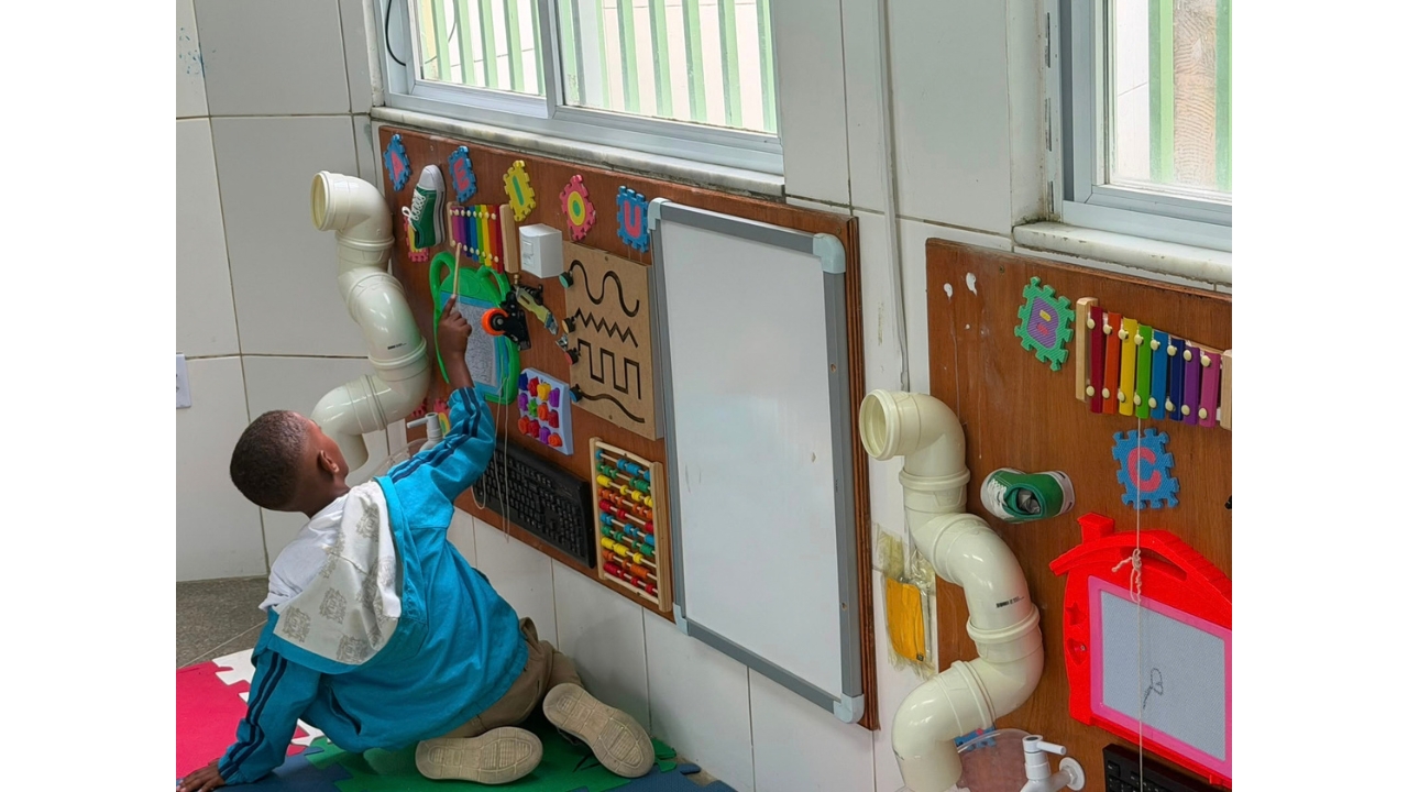 Espaço interno destinado a atividades sensoriais, onde uma criança ajoelhada interage com um painel fixado à parede. O painel reúne ábaco, formas, letras, tubos, instrumentos coloridos e objetos de manipulação. Ao fundo, há janelas e paredes revestidas de azulejos brancos.