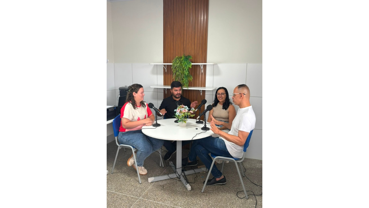 Sala de reunião com quatro adultos sentados ao redor de uma mesa redonda, participando de uma conversa com microfones. Duas mulheres e dois homens gesticulam e observam uns aos outros. Ao centro da mesa, há um vaso com flores; ao fundo, aparecem prateleiras e uma parede com revestimento de madeira.