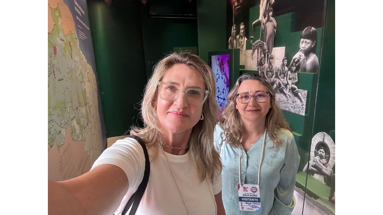 Em uma exposição, duas mulheres de óculos posam para uma foto estilo selfie; uma veste blusa branca de mangas curtas e a outra, blusa azul-clara de mangas compridas com crachá de visitante. Ao fundo, há mapas, fotografias em preto e branco e painéis de temática cultural, com retratos de povos indígenas e um fundo verde.
