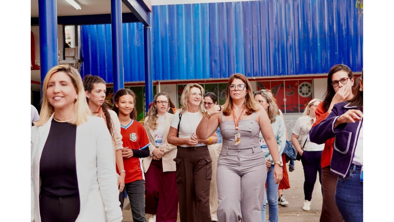 Área externa de uma escola, com ao menos 12 pessoas caminhando em grupo, todas mulheres. Elas usam roupas casuais e crachás, e uma estudante veste uma camiseta vermelha e calça jeans. Ao fundo, há uma estrutura azul e a fachada escolar.