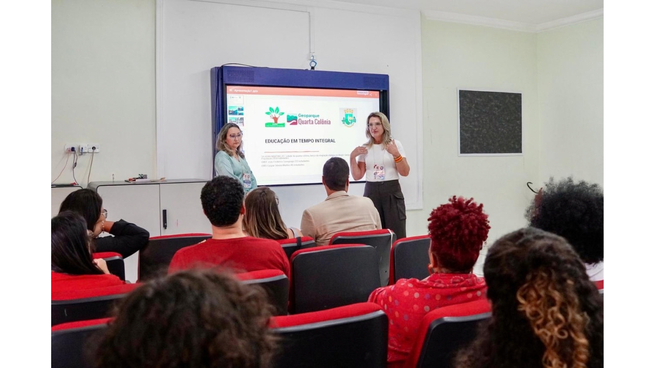 Sala de apresentação de paredes claras e público sentado em cadeiras com estofado vermelho. Duas mulheres estão em pé diante de uma tela de projeção; uma delas gesticula enquanto apresenta o conteúdo “Geoparque Quarta Colônia - Educação em Tempo Integral”.v