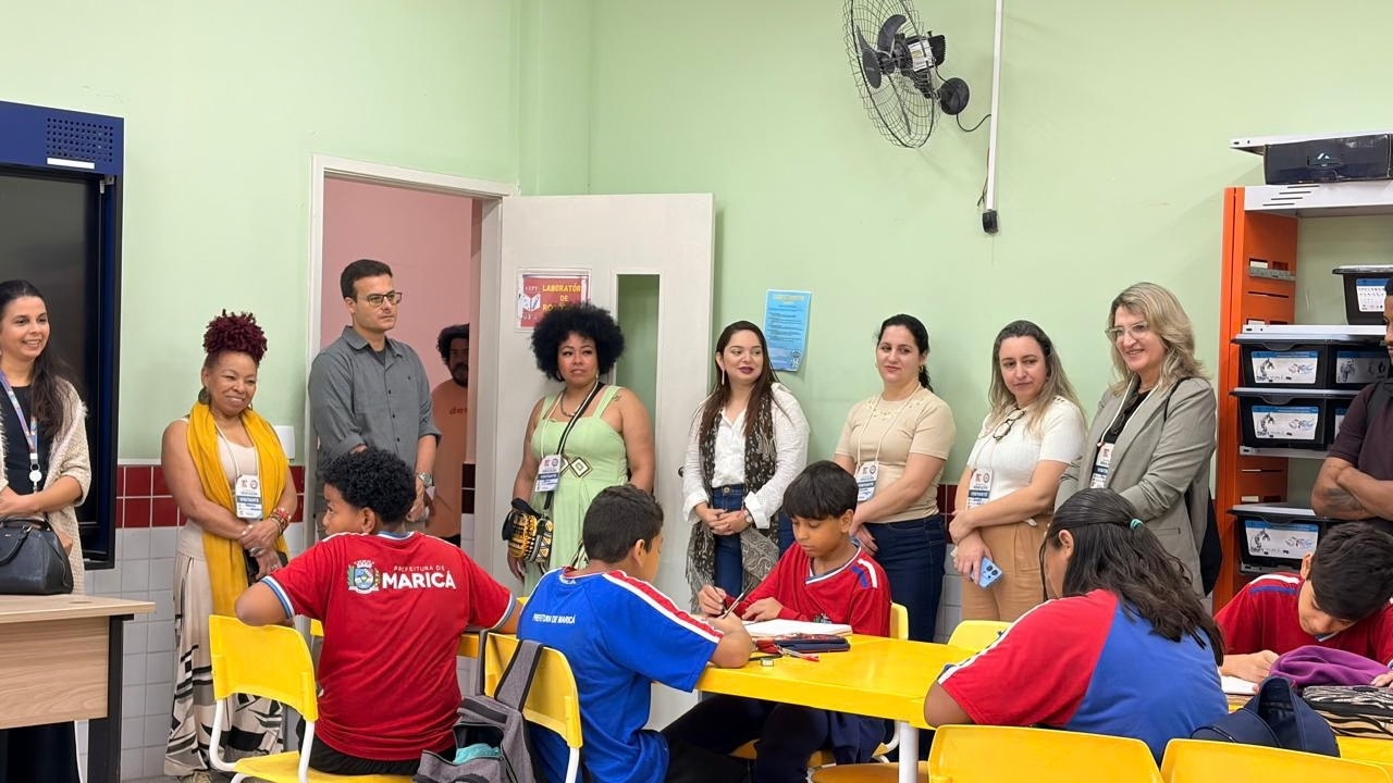 Sala de aula com estudantes sentadas(os) em mesas e cadeiras amarelas, realizando atividades. Um grupo multirracial de oito adultos com crachás observa as(os) alunas(os), que vestem roupas vermelhas e azuis com a inscrição "Prefeitura de Maricá" nas costas, juntamente com o brasão da cidade. Ao fundo, há armários, estantes e paredes verdes. A porta branca da sala está aberta e há um vestilador fixado na parede.